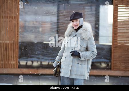 Porträt stilvolle reife Frau steht draußen im Schnee, trägt modisches mehrlagiges Winteroutfit, graue Jacke mit Taschen, Kapuze und Kappe. Älterer F Stockfoto