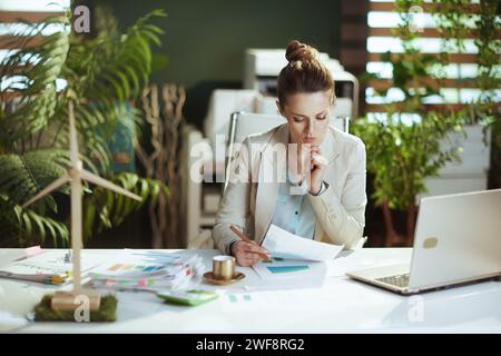 Nachdenkliche, moderne Geschäftsfrau mittleren Alters in einem leichten Business-Anzug in einem modernen grünen Büro mit Dokumenten und Laptop. Stockfoto