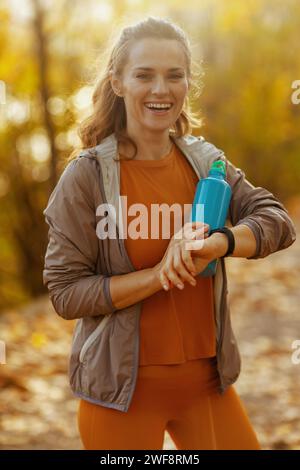 Hallo Herbst. Glückliche moderne 40-jährige Frau in Fitness-Kleidung im Park mit einer Flasche Wassersport-Tracker. Stockfoto
