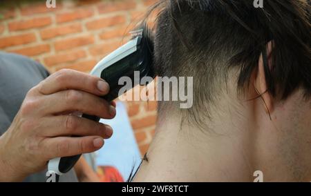 Die Hand eines Mannes hält einen Haarschneider mit einer Düse und schneidet einem jungen Mann den Hinterkopf, Nahaufnahme, Männer in der Familie kümmern sich um einander Stockfoto