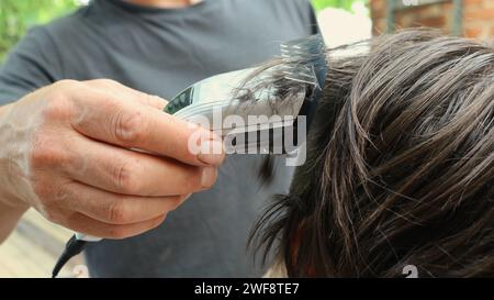 Die männliche Hand eines Friseurs hält eine Haarmaschine nahe dem Kopf einer langhaarigen Person und schneidet langes Haar mit einem Trimmer mit Aufsatz Stockfoto