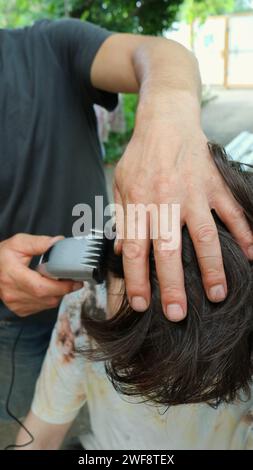 Ein Mann schneidet die langen Haare eines Teenagers auf dem Hof eines Privathauses mit einem Haarschneider, Nahaufnahme, vertikalem Bild, männlichen Familienbeziehungen Stockfoto