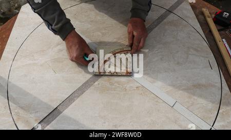 Die Hände des Flieseners legen ein quadratisches Stück Material mit einem Blumenmuster auf die glatte Oberfläche einer großen hellen Fliese und bilden ein Mosaik Stockfoto