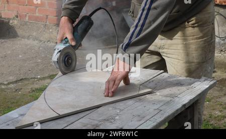 Ein Meister sägt ein großes Stück Fliese entlang des Umfangs mit einer Gehrungssäge in einer Werkstatt im Freien ab und schneidet eine runde Form aus Keramikfliesen aus Stockfoto