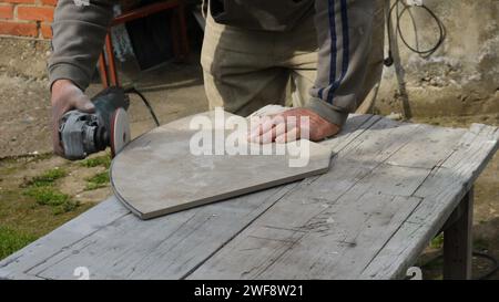 Die Kanten einer Fliese mit einem Schleifgerät mit Schleifaufsatz schleifen, hält der Arbeiter eine große geschnittene Fliese mit der Hand auf eine Holzarbeitsfläche Stockfoto