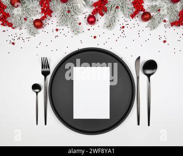Weihnachtskomposition, schwarzer Teller und Besteck, weiße Karte, Tannenzweige, rote Beeren, Kugeln auf weißem Hintergrund. Stockfoto