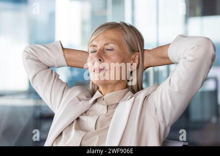 Nahaufnahme einer älteren, grauhaarigen Geschäftsfrau in einem Anzug, die auf einem Stuhl im Büro sitzt und sich ausruht, Hände hinter dem Kopf und Augen geschlossen. Stockfoto