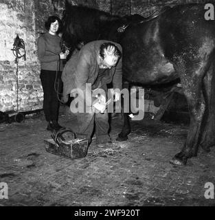 1970er Jahre, historisch, ein älterer Schmied oder Farmer, bei der Arbeit in Stallungen, bückte sich, während er den Hu eines Pferdes schuf, ein Stallmädchen, das den Pferdekopf hält, England, Großbritannien. Stockfoto