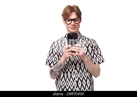 Junger gutaussehender kaukasier mit rötlich goldenen Haaren in einem schwarz-weißen Sommerhemd macht ein Foto auf einem Smartphone Stockfoto