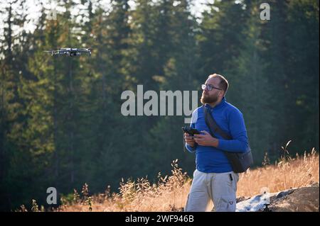 Professioneller Fotograf, der Fotos von der Drohne macht. Junger Mann, der Luftaufnahmen in den Bergen macht. Mann, der seine Drohne auf dem Hintergrund des Bergwaldes kontrolliert. Stockfoto