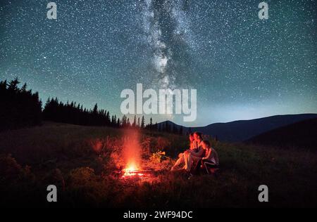 Gemütliche Nacht mit Familie in den Bergen. Junge Familie sitzt am Lagerfeuer in sternenklarer Nacht. Heller Blick auf die Milchstraße über die Hügel. Stockfoto