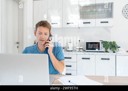 Fröhlicher junger Geschäftsmann, der telefoniert, während er zu Hause am Laptop arbeitet. Männlicher professioneller Broker-Investor, der online an der Börse arbeitet Stockfoto