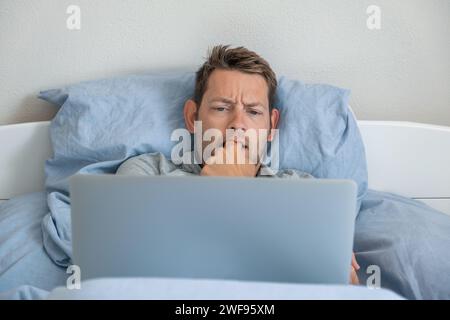 Zufriedener junger Geschäftsmann freiberuflich am Laptop, der zu Hause im Bett liegt. Männlicher professioneller Händler Investor, der online an der Börse arbeitet und Stockfoto