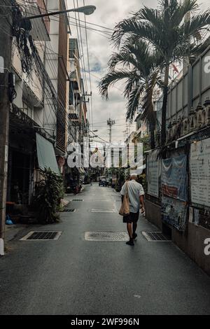 Eine einsame Person läuft von der Kamera in einer ruhigen, engen Gasse, die von Gebäuden und Palmen gesäumt ist, unter einem bewölkten Himmel. Stockfoto