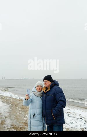 Porträt eines älteren Ehepaares, das einen Winterspaziergang am Strand macht und ein Selfie macht. Vertikaler Rahmen. Stockfoto