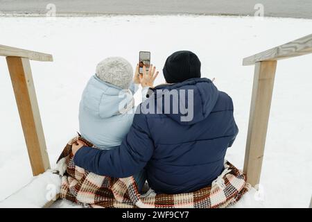 Glückliches älteres Paar, das im Winter auf der Treppe am Meer sitzt und ein Selfie macht. Stockfoto