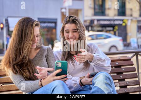 Zwei Frauen lachen, während sie auf ihre Handys schauen, die auf einer Bank sitzen. Ein paar Frauen, die nach sozialen Netzwerken süchtig sind. Stockfoto