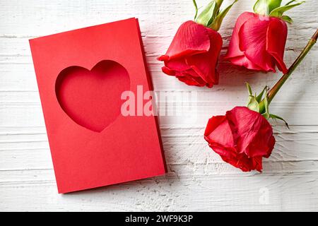 Rote Rosen und herzförmige Grußkarte auf weißem hölzernem Hintergrund, Blick von oben Stockfoto