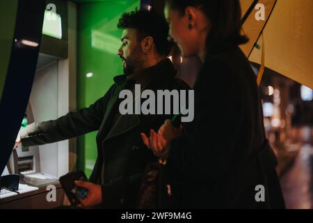 Ein Paar, das an einem regnerischen Tag einen Geldautomaten benutzt. Der Mann hebt Geld ab, während er einen Schirm hält, und sie unterhalten sich. Stockfoto