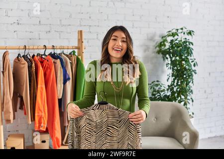 Begeisterte asiatische Stylistin mit maßgeschneiderter Bluse mit Tiermuster, die im Modestudio vor der Kamera lächelt Stockfoto