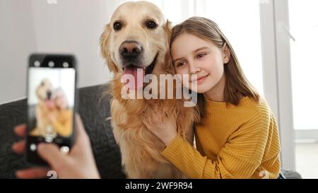 Mädchenhand mit Smartphone, das Foto von Golden Retriever Hund und Kind zu Hause macht. Frau Mutter fotografiert Haustier Hund und Tochter mit Handykamera Stockfoto