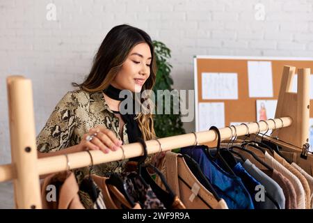 Fröhlicher asiatischer Designer, der in der Nähe des Regals lächelt, mit stilvoller, maßgeschneiderter Kleidung in einem persönlichen Modeatelier Stockfoto