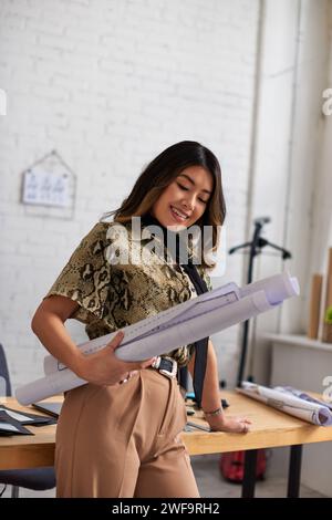 Junger und glücklicher asiatischer Designer, der mit Nähmustern in der Nähe des Schreibtisches im Modeatelier steht Stockfoto