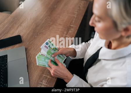 Blick von oben auf eine glückliche reiche Geschäftsfrau, die einen Haufen Euro-Dollar zählte und einen Schreibtisch in eleganter Kleidung trug. Nahaufnahme von weiblichen Händen, die hundert halten Stockfoto