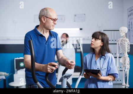 Physiotherapie beim Pedalfahren mit dem Heimtrainer mit Hilfe einer in Peelings gekleideten Krankenschwester. Medizinischer Assistent hält Tablette, um medizinische Ergebnisse an alten Mann weiterzugeben. Stockfoto