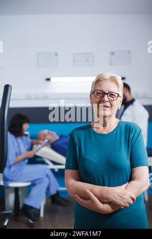 Eine alte Frau im Porträt wartet auf körperliche Behandlung mit überkreuzten Armen. Ältere Patientin mit Muskelverletzung macht sich bereit für die Beinphysiotherapie auf dem Heimtrainer, während sie in die Kamera schaut. Stockfoto