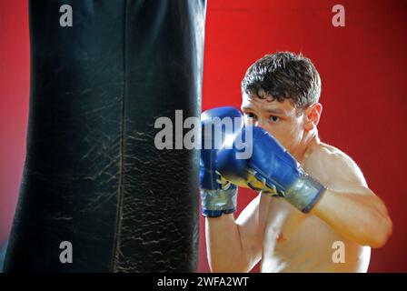 Ein fokussierter Boxer mit blauen Boxhandschuhen macht sich bereit, eine schwere Tasche zu treffen Stockfoto