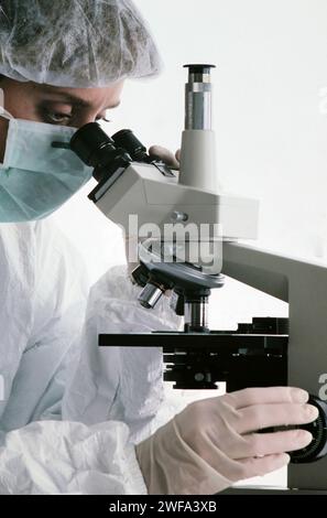 Ein fokussierter Wissenschaftler, der in einen Labormantel und eine Schutzmaske gekleidet ist, untersucht eine Probe sorgfältig durch die Linse eines modernen Mikroskops in einer sauberen Arbeit Stockfoto