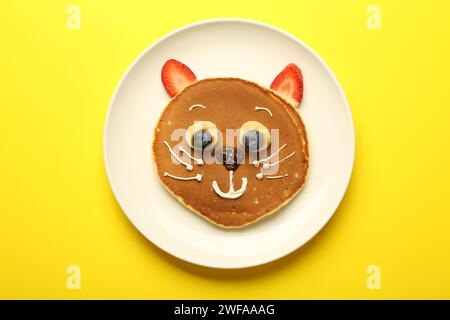 Kreatives Servieren für Kinder. Teller mit niedlicher Katze aus Pfannkuchen, Beeren, Creme, Banane und Schokoladenpaste auf gelbem Hintergrund, Draufsicht Stockfoto