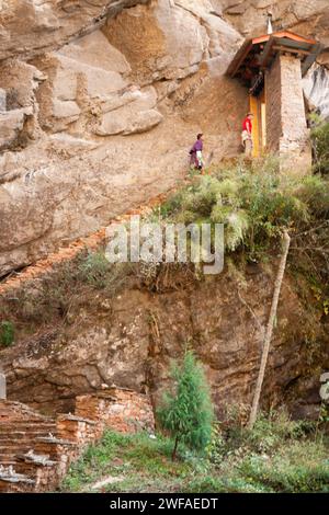 Bhutanischer Mann mit traditionellem Ghos und weibliche Touristen klettern die steilen Treppen auf dem felsigen Pfad zum Kloster Tiger's Nest, bereit, durch A d zu gehen Stockfoto