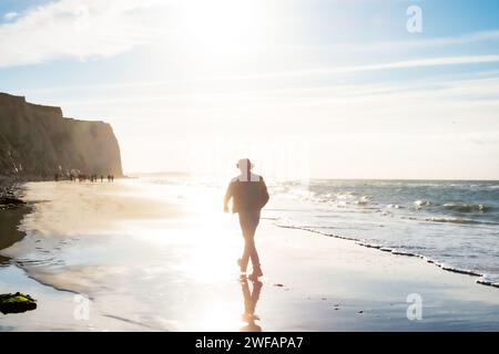 Das Bild zeigt eine Einzelperson, die sich auf einen gemütlichen Spaziergang entlang der Küste aufmacht, während die Morgensonne ein diffuses Licht über die Szene wirft. Dieses Mal erscheint die Person näher und prominenter und deutet auf eine Erzählung der Interaktion mit der Umwelt hin. Das Morgenlicht macht die Konturen der Klippen und der Figur weich und schafft eine traumhafte Qualität. Kleine Wellen brechen sanft am Ufer, während der nasse Sand die Figur und das glitzernde Sonnenlicht reflektiert und das Gefühl der Ruhe am frühen Morgen verstärkt. Küstenfahrt bei Dawn. Hochwertige Fotos Stockfoto