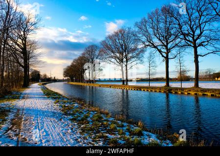 Dieses fesselnde Bild zeigt einen schneebedeckten Pfad entlang eines ruhigen Kanals, flankiert von majestätischen, blattlosen Bäumen vor einem hellen Winterhimmel. Die untergehende Sonne wirft ein sanftes Licht und erzeugt einen lebhaften Kontrast zwischen dem Blau des Himmels und der goldenen Reflexion auf der Wasseroberfläche. Der schneebedeckte Pfad mit Spuren führt den Blick zum Horizont, während der Kanal eine parallele Reise entlang bietet. Das Vorhandensein von Wolken verleiht dem Himmel eine dynamische Textur und ergänzt die Stille des Kanals. Frischer Wintertag am Kanal. Hochwertige Fotos Stockfoto