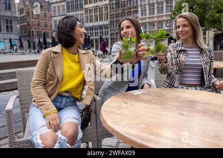 Dieses Bild ist lebendig mit der Lebendigkeit des Stadtlebens, während eine Gruppe von Freunden in einem Café im Freien ihre Gläser hebt. Die zentrale Figur wird mitten im Lachen gefangen und greift mit ihrem Getränk zur Kamera, umgeben von Freunden, die ihre Freude teilen. Die Umgebung ist ein eleganter Stadtplatz mit historischen Gebäuden im Hintergrund, die die Themen betonen. Der bewölkte Himmel wirft ein natürliches, diffuses Licht über die Szene und verstärkt die Spontaneität und Wärme des Augenblicks. Urban Toast: Eine lebhafte Begegnung im City Cafe. Hochwertige Fotos Stockfoto