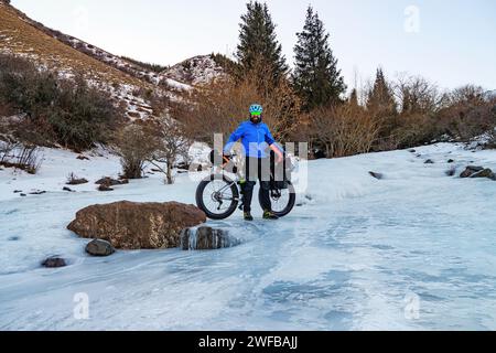 Ein bärtiger Mann reist im Winter mit einem fetten Fahrrad in die Berge. Ein Mann in blauer Jacke und Helm steht mit einem Fahrrad auf dem Eis eines gefrorenen Flusses. Stockfoto