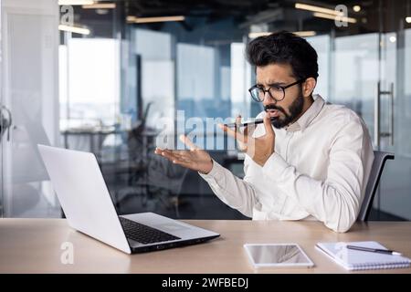 Ernsthafter junger indischer Geschäftsmann sitzt im Büro am Schreibtisch und spricht emotional am Telefon über den Lautsprecher, verwendet Sprachsuche und nimmt das Gespräch auf. Stockfoto