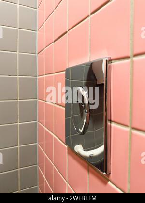 Ein Chrom glänzender WC-Spülknopf an der pinkfarbenen Wand mit kontrastierenden grauen Wandfliesen auf der gegenüberliegenden Seite Stockfoto