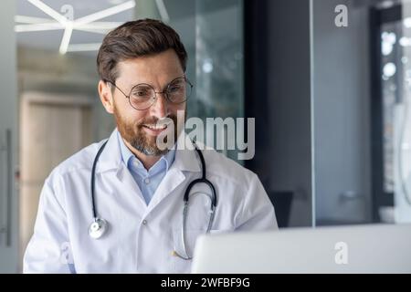 Nahaufnahme eines jungen, lächelnden männlichen Arztes, der im Büro sitzt und auf einem Laptop bei einem Videoanruf spricht. Stockfoto