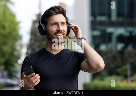 Nahaufnahme eines jungen Athletikers, der auf einer Stadtstraße mit Kopfhörern steht, ein Telefon in der Hand hält und mit einem Lächeln zur Seite blickt. Stockfoto