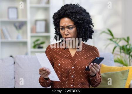 Verwirrte junge Frau mit Brille, die Smartphone und Papier hält, Bedenken oder Zweifel zum Ausdruck bringt, Inneneinrichtung mit Sofa und Bücherregal. Stockfoto
