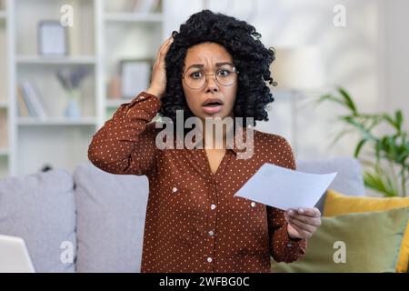 Besorgte junge Frau mit Brille, die ein Dokument hält, Stress und Verwirrung zu Hause ausdrückt, Konzept der finanziellen oder persönlichen Krise. Stockfoto