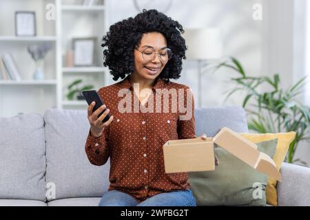 Fröhliche Frau mit lockigem Haar mit Smartphone und offenem Paketkasten, während sie auf einer Couch in einem gemütlichen Wohnzimmer sitzt. Stockfoto