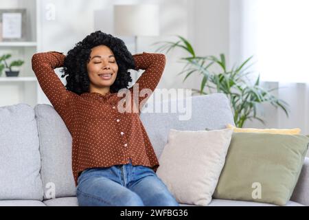 Entspannte afroamerikanische Frau genießt einen friedlichen Moment, liegend auf einer Couch zu Hause, die Augen geschlossen mit einem zufriedenen Lächeln. Stockfoto