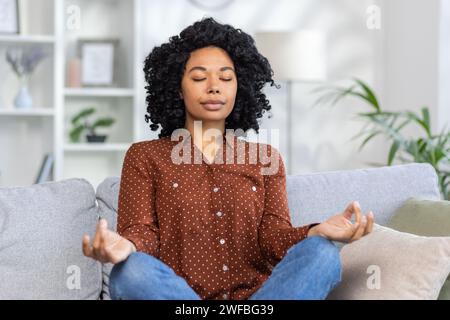 Ruhige Frau, die auf der Couch meditiert, Augen geschlossen in friedlicher Atmosphäre, Achtsamkeit und Entspannungskonzept Stockfoto