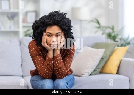 Verärgerte junge afroamerikanische Frau, die mit dem Kopf nach unten auf dem Sofa sitzt. Sie legte ihren Kopf auf ihre Hände und schaute nachdenklich zur Seite. Stockfoto