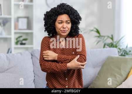 Nahaufnahme einer traurigen Afroamerikanerin, die zu Hause auf dem Sofa sitzt und ihren Ellenbogen mit der Hand hält, vor Schmerzen zuwinkelt und an einer Verstauchung leidet. Stockfoto