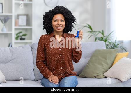 Porträt einer jungen, lächelnden Afroamerikanerin, die zu Hause auf der Couch vor der Kamera sitzt und Asthmaspray zeigt. Stockfoto